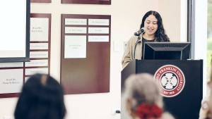 A young woman stands at a podium. The heads of audience members are visible in the foreground of the photo.