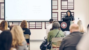 A young woman speaks at a podium in front of a small crowd, while an image of a drawing of a white medical coat is projected onto a screen.