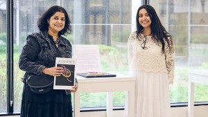 A young woman and her mother stand on either side of a clear display case containing a 3D artwork, holding a copy of the magazine Synapses with that artwork on the cover.