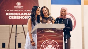 Three students stand onstage behind a podium, with a banner reading "Chicago Medical School Aesculapian Ceremony" in the background.