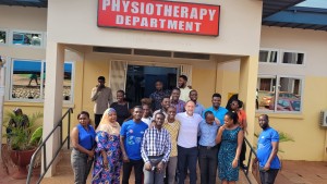 A man stands surrounded by a large group of people, in front of a yellow building with a red sign that reads "Physiotherapy Department"