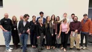 A diverse group of fifteen people stand smiling against a blank wall.