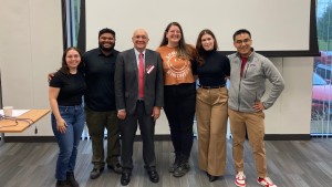 A man in a suit and a red tie stands with a group of students in front of a projector screen