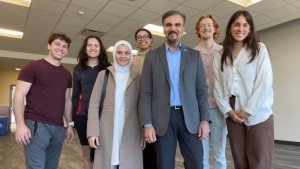 A man in a suit jacket and blue shirt smiles at the camera, surrounded by six smiling medical students
