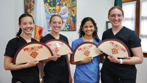Four young women wearing scrubs hold up paper fans that say Veracruz