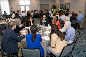 A diverse group of people sits around tables in a conference room, engaged in discussion. The atmosphere is collaborative and focused, with notes and coffee cups visible.