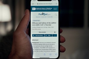 A smartphone displays a PubMed article titled "Efficacy and Safety of the mRNA-1273 SARS-CoV-2 Vaccine." A hand holds the phone in focus.
