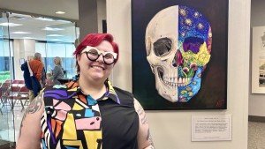 A young woman in a colorful shirt stands next to an artwork of a human skull. The left half of the skull is realistically painted, while the right half is decorated with colorful paintings of flowers, wheat fields, the night sky, and water lilies.