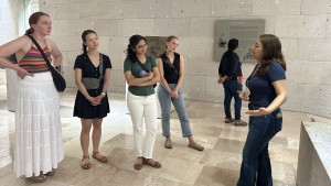 Five students stand in a building with a stone floor, listening to a woman speaking to them