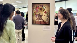A smiling woman stands next to an artwork depicting a woman smelling a rose, while flowers cover the top of her head. Another woman is standing to the side looking at the artwork.