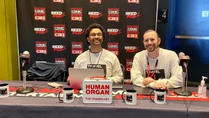 Two men sit at a table, with the backdrop behind them showing that they are at the Chicago Comic and Entertainment Expo. On the table, there are several coffee mugs that say "Travel Medicine" and there is a white cooler that says "Human Organ for Transplant" in large red letters.