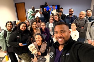 Group of diverse individuals smiling in a classroom. One person in front takes a selfie. The mood is cheerful and collaborative. Tables and chairs are visible.