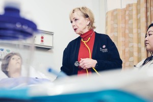A nurse in a red shirt and black cardigan with a stethoscope examines a medical mannequin in a hospital room, conveying a learning environment.