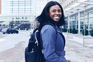 A student in a blue coat stands outdoors with a backpack, smiling towards the camera.
