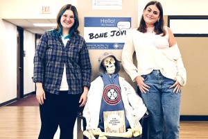 Two smiling individuals pose with a skeleton named "Bone Jovi" wearing a hat and a library vest, sitting in a chair, creating a lighthearted tone.