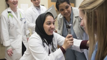 A female student using an ophthalmoscope to examine a patient's eye, surrounded by smiling colleagues in lab coats. 
