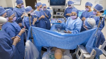 A group of students in blue scrubs and masks surround a mannequin in an operating room, focusing on a procedure. 