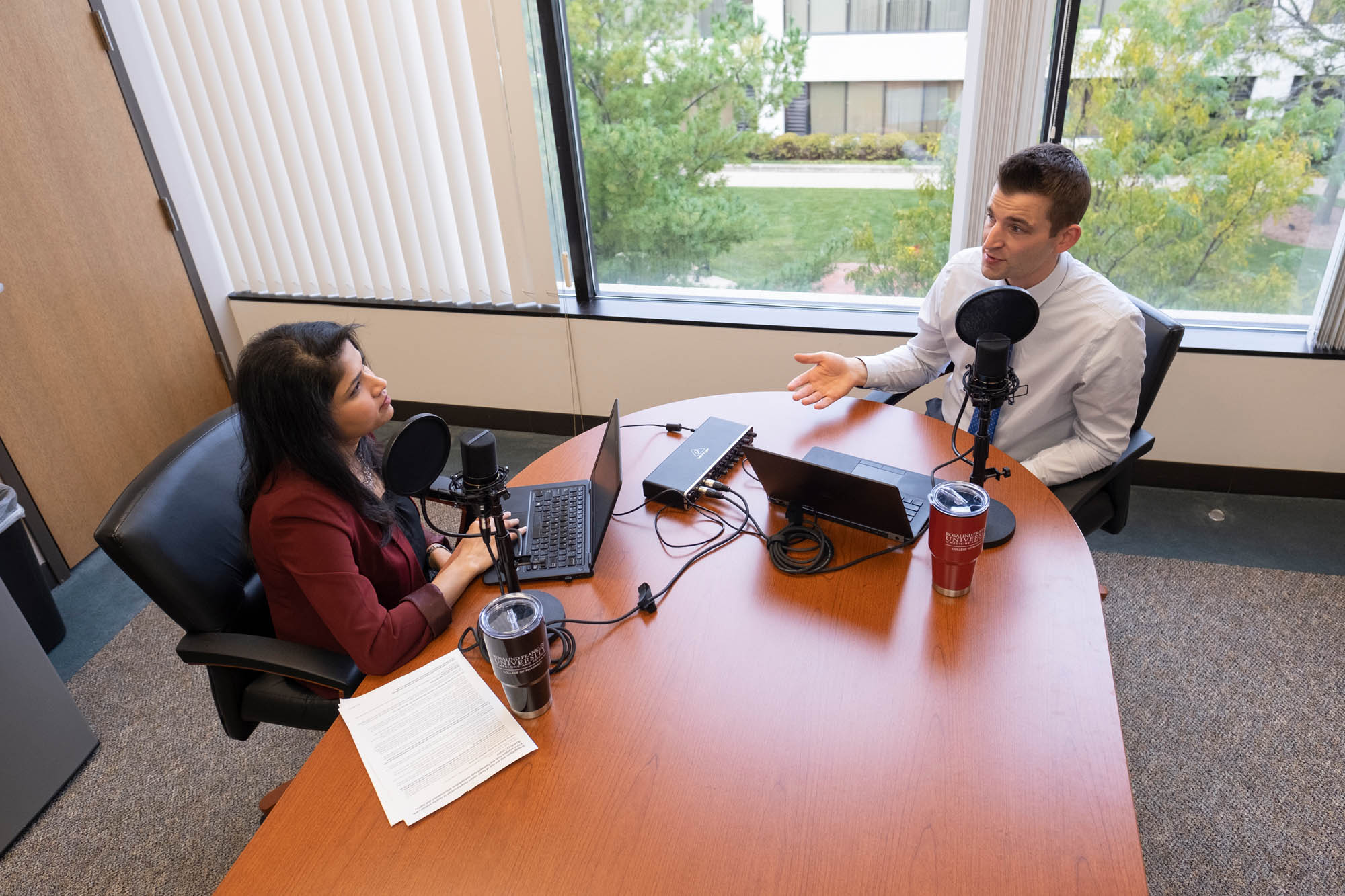 Sean P. Kane, PharmD, BCPS and Khyati S. Patel, PharmD, BCACP sit at a table with a laptop, engaged in a discussion about their podcast HelixTalk.