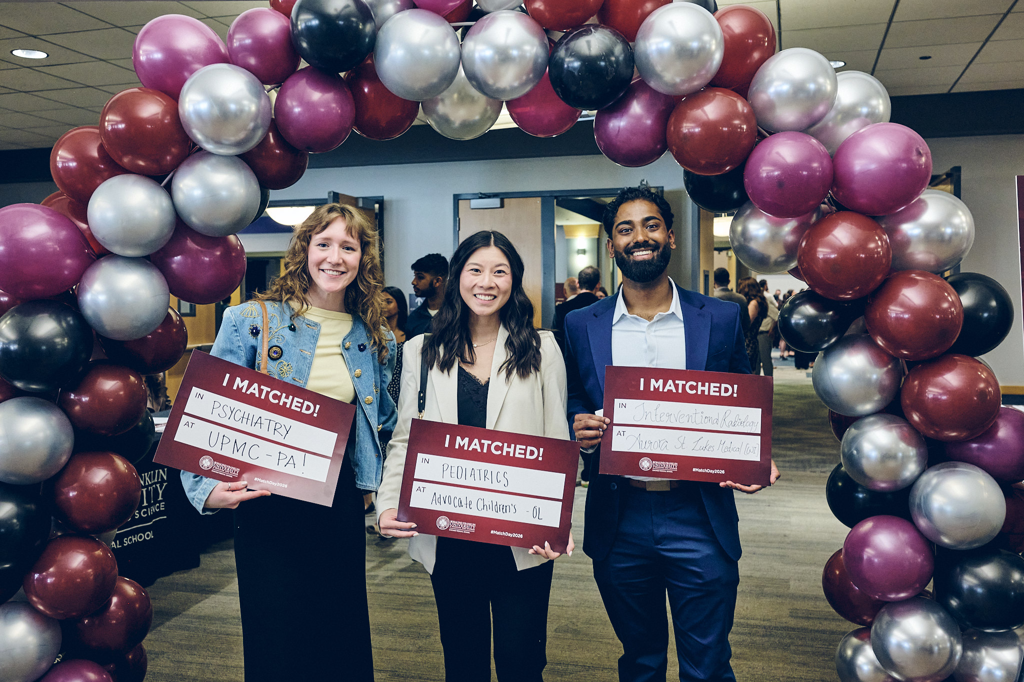 Three smiling medical students stand under a balloon arch holding signs reading "I matched!"