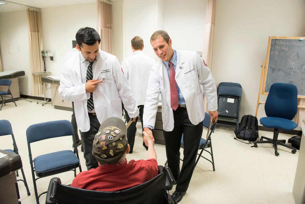 Two medical students wearing white coats speak to a man in a wheelchair