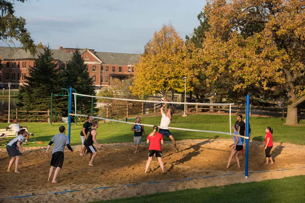 A group of male and female students play volleyball on an outdoor, sand-filled court, with trees and a brick building in the background.